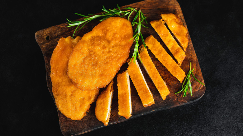 Breaded chicken cutlets on a wooden cutting board with sprigs of rosemary