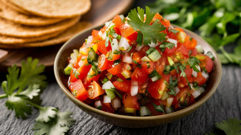 A bowl of fresh salsa with cilantro and tortillas in the background