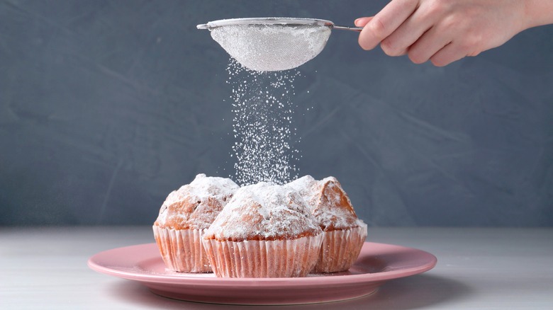 person shaking powered sugar in sieve over a muffins