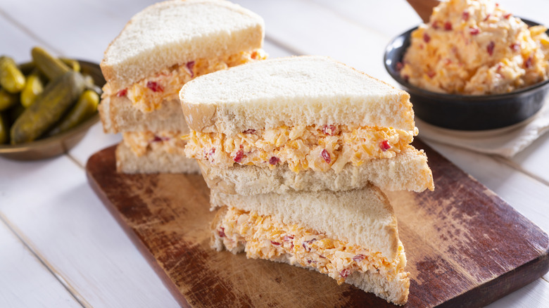 Pimento cheese sandwiches with white bread, cut diagonally, near bowls of pimento cheese spread and pickles