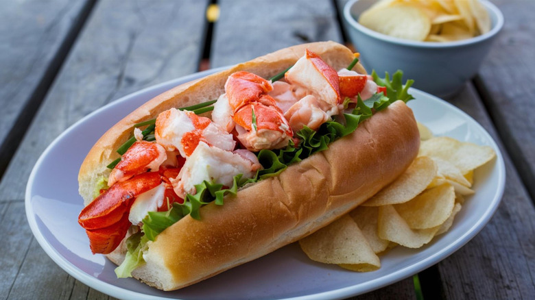 Maine lobster roll with lobster chunks on lettuce on a roll, next to potato chips on a plate