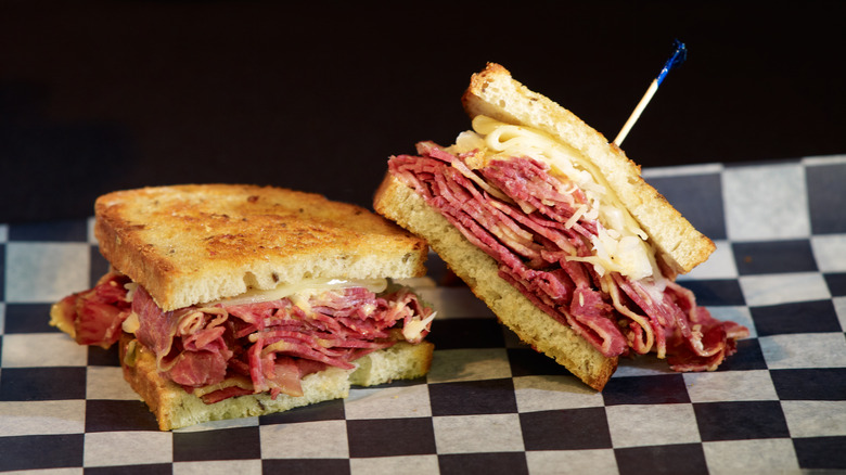 Two halves of a reuben sandwich on checkerboard paper, one half has a toothpick with blue cellophane on it