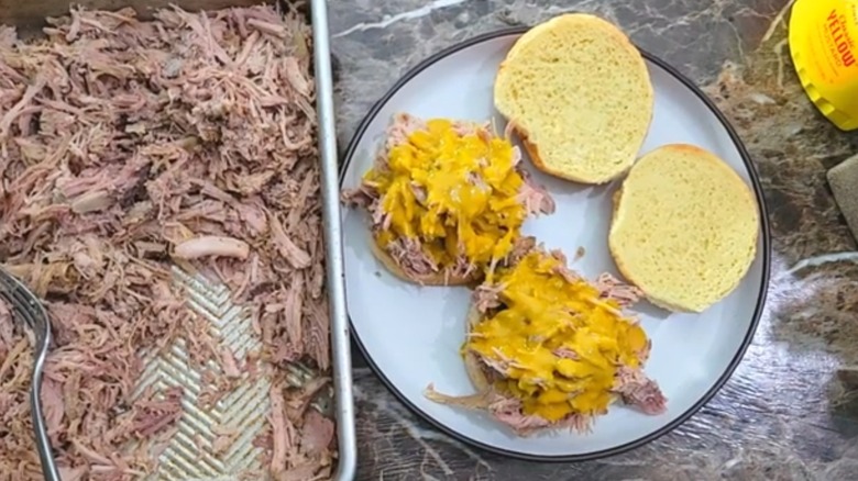 Two pulled pork sandwiches with homemade Carolina gold sauce, top bun halves on the side, on white plate next to pan of pulled pork