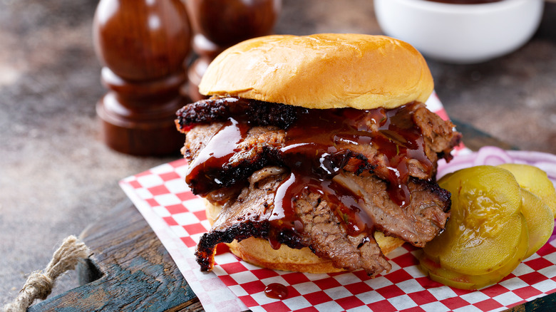 Brisket on a bun, dripping with BBQ sauce, on red checkerboard paper next to pickles and red onions