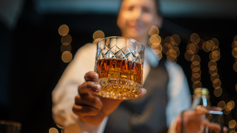 A bartender handing a glass of whiskey toward the camera