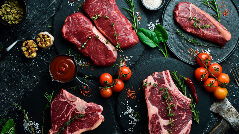 Several raw steak varieties arranged with tomatoes, herbs, and garlic.