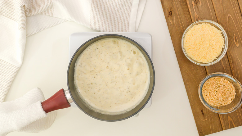 Saucepan of Alfredo sauce next to cheese and breadcrumbs