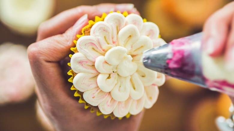 Hands doing intricate floral piping on a cupcake