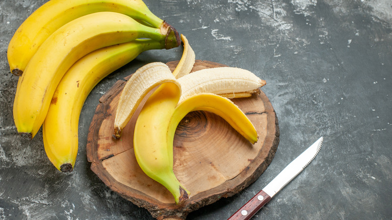 A peeled banana on a cutting board with a bunch of bananas and a knife