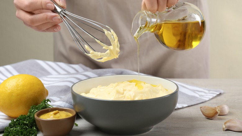 Hand pouring oil into bowl of mayo with whisk