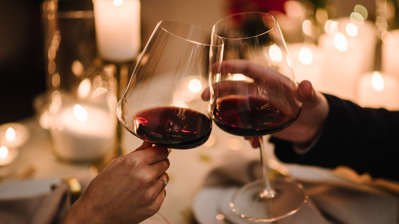 Two people's hands toast with red wine glasses over a candlelit restaurant table