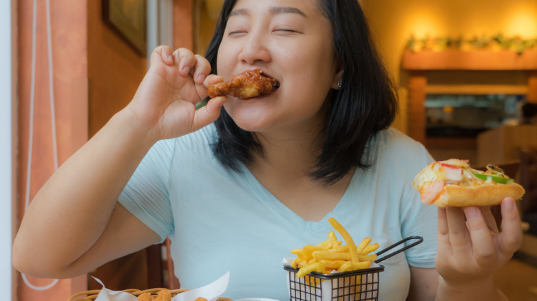 A smiling woman eats a chicken wing while holding a piece of pizza in her other hand.