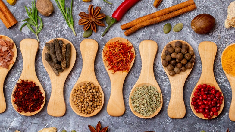 A variety of whole spices displayed on a row of wooden spoons pointed in alternating directions with loose whole spices scattered around them.