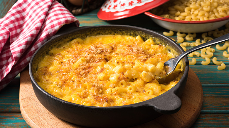 Serving dish is filled with mac and cheese with a checkered cloth and uncooked noodles in background.