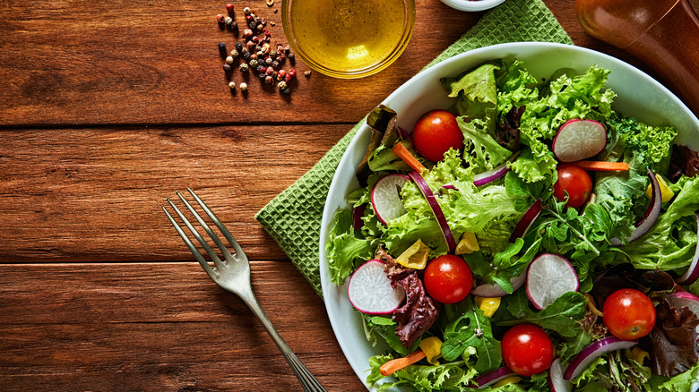 A green salad in a white bowl sitting on a wooden table next to dressing and a fork