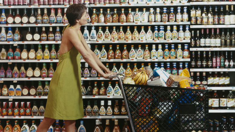 A woman with brown hair wearing a green dress pushing a black shopping cart filled with various groceries past a grocery aisle with shelves full of various salad dressings