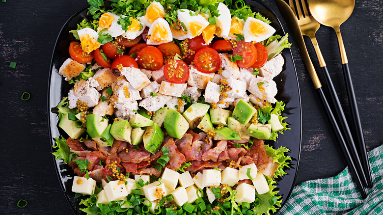 Assembled cobb salad on a black background with gold and black silverware