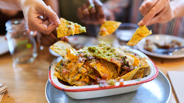 Several hands grabbing chips from a bowl of nachos.