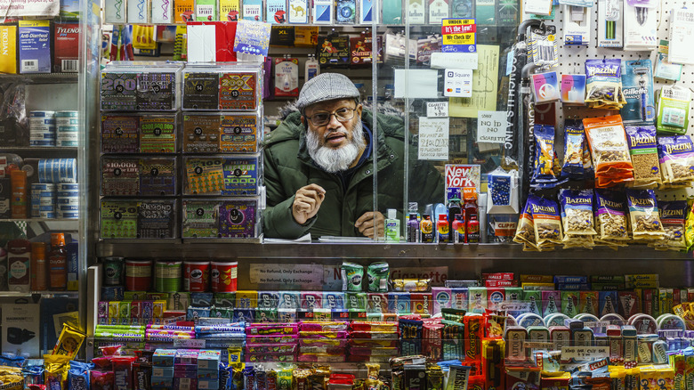 A man behind a packed convenience store counter looking into the camera