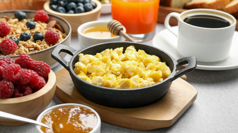 A small cast iron pan filled with yellow scrambled eggs sitting on a wooden platter, with a cup of coffee, a glass of orange-colored juice, a wooden bowl filled with blueberries, a white ceramic bowl filled with granola and blueberries and raspberries, and a white dish of honey with a wooden honey dipper in the background. In the foreground is a wooden bowl filled with raspberries and a small white bowl filled with a brown-colored jam.