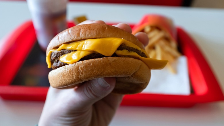 A hand holding a burger, with fries and a drink in the background