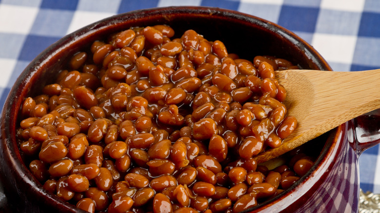A wooden spoon spooning out a helping of glossy, sticky baked beans from a crockery pot sitting on a gingham tablecloth.