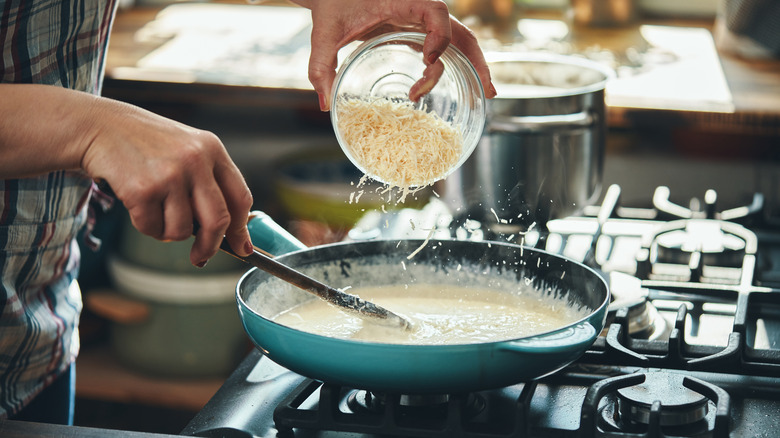 Closeup of hands sprinkling grated parmesan cheese while stirring a pan of alfredo sauce