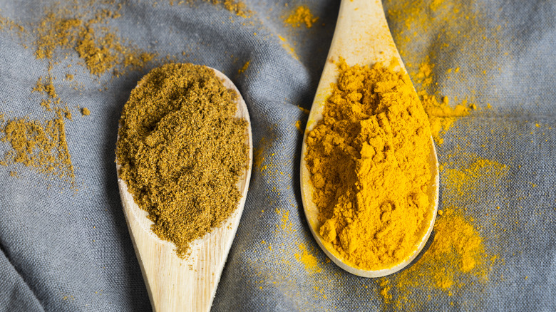 Two wooden spoons each holding different spices, one brown and one yellow in color, on a gray tablecloth