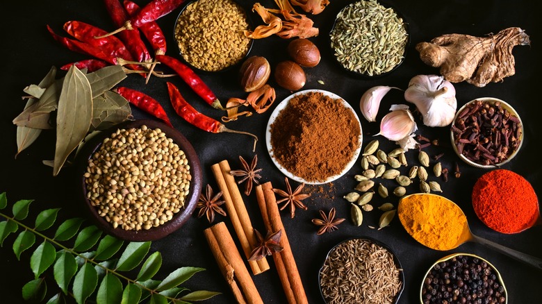 A variety of spices including cinnamon sticks, ginger, garlic cloves, chili peppers, bay leaves, and various powders and seeds, on a black tabletop