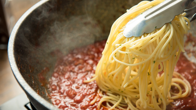 Tongs putting long pasta into a pan of tomato sauce