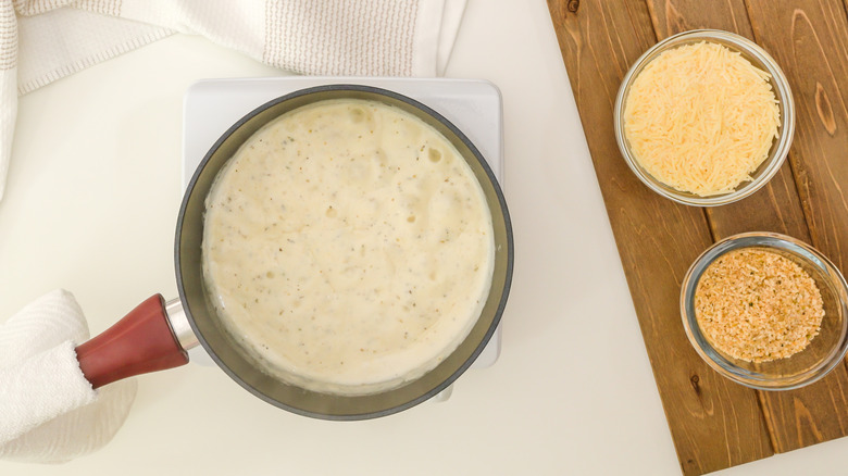 Saucepan of Alfredo sauce next to cheese and breadcrumbs