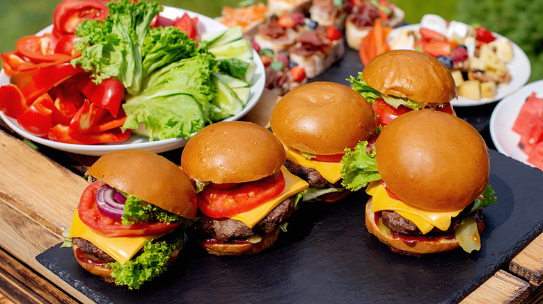 Several burgers arranged beside a bowl of lettuce and tomatoes at a cookout.