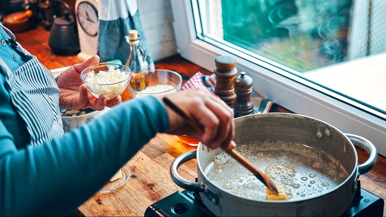 Home cook melting butter in a pan while holding a small glass cup of onions