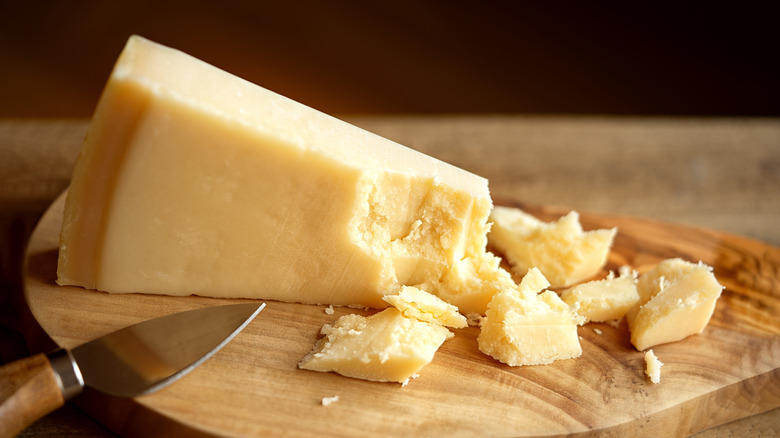 A wedge of cheese like Grana Padano is partially crumbled on a wooden cutting board.