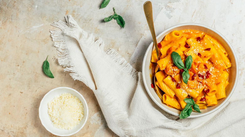 A bowl of rigatoni in a bright orange-yellow sauce is near a smaller bowl of grated cheese like Grana Padano.