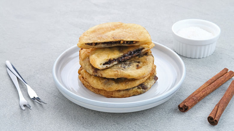 A plate of homemade hotteok is displayed with cinnamon sticks and a bowl of sugar.