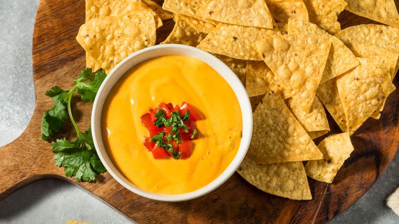 A bowl of queso with tomatoes and cilantro on top next to tortilla chips on wooden cutting board