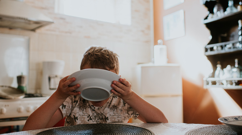 A kid sips from a bowl at a kitchen table.