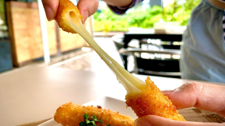 Hands holding a mozzarella cheese stick with one end in each hand and melted cheese stretching between the two ends