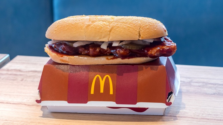 A McRib sandwich sits on a fast food carton on a wooden table.