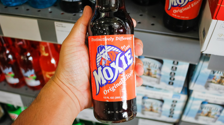 A hand holding a bottle of Moxie Original Elixir soda
