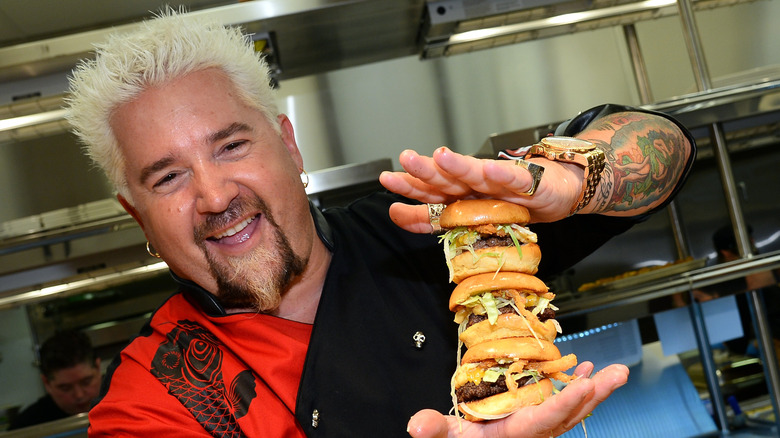 A headshot of Guy Fieri holding a stack of burgers