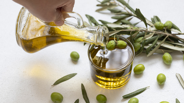 A hand pours olive oil from a carafe into a clear glass surrounded by olives