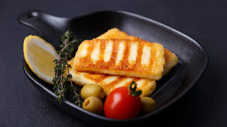 Grilled halloumi is placed in a black skillet with tomato, olives, thyme, and lemon.