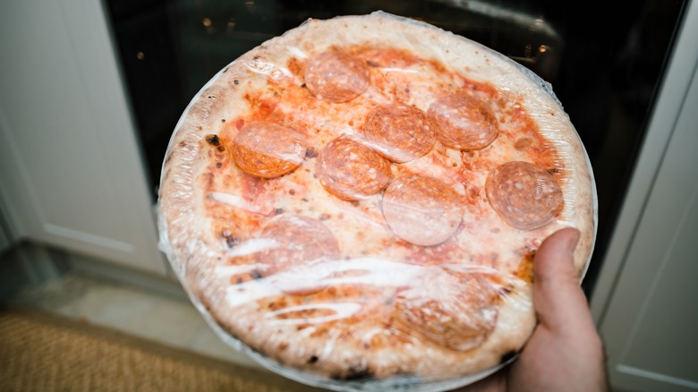 Person holding a plastic-wrapped frozen pepperoni pizza