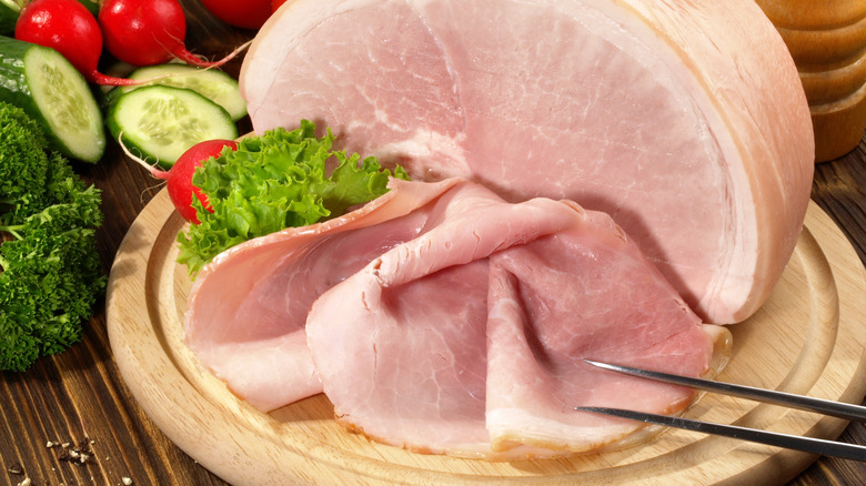 Sliced, cooked ham on a cutting board with fresh vegetables