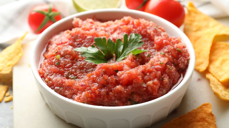 salsa in white bowl with chips and tomatoes and lime in background