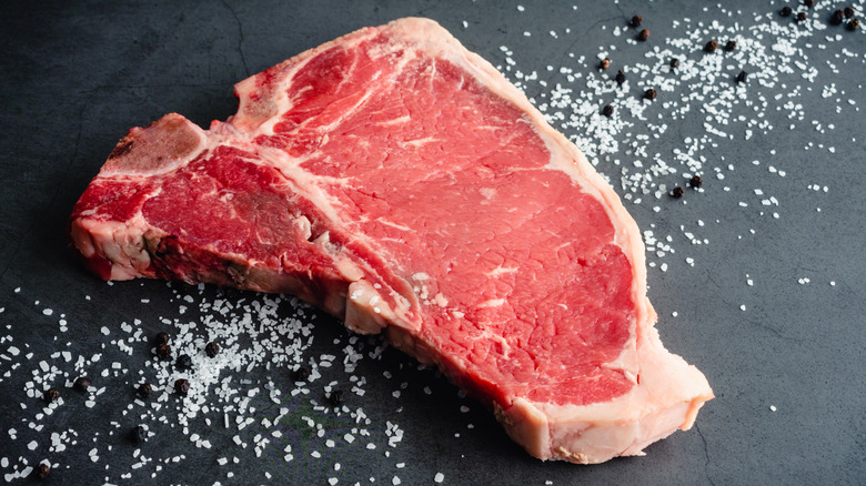 Raw t-bone steak sits on a black countertop surrounded by salt and peppercorns