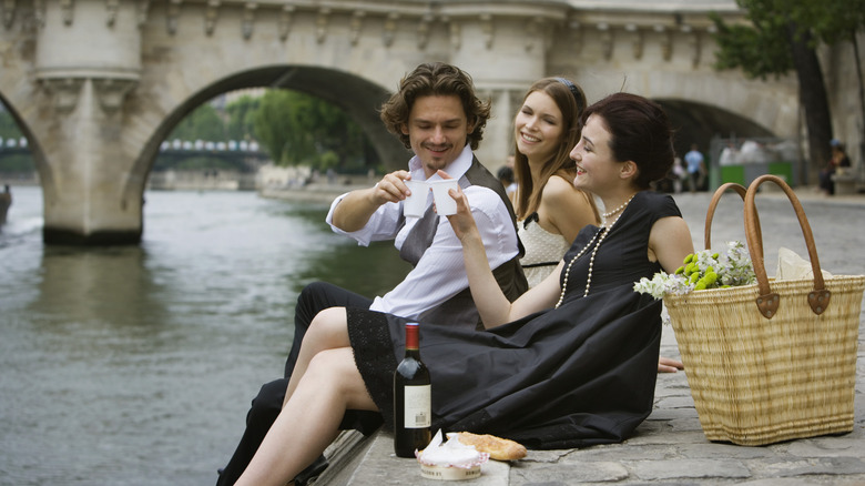 Three swankily dressed people having wine in plastic cups, Camembert, and baguette on the banks of the Seine, by the Pont Neuf