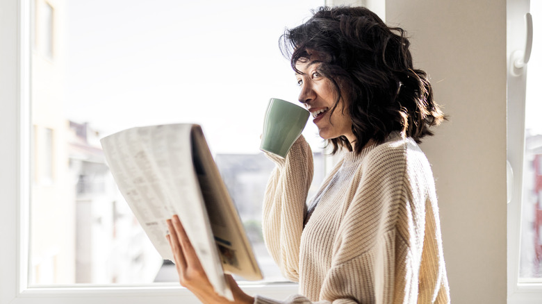 A person reads a newspaper and sips from a coffee cup.